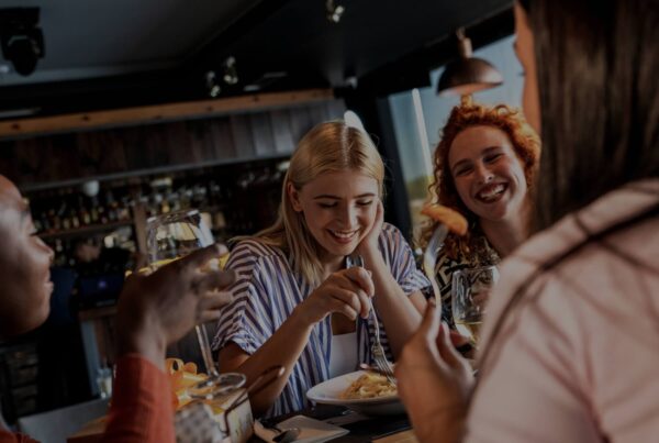 Four people sit around a table at a restaurant, laughing and enjoying food together. The warm, friendly atmosphere hints at the exciting new LAX shops & eateries, with plates and drinks adding to the inviting scene.