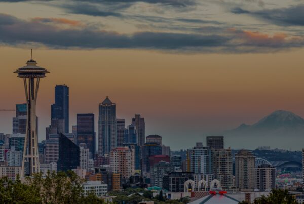 Seattle skyline at sunset with the Space Needle in the foreground, tall buildings downtown, and Mount Rainier faintly visible—a stunning scene from one of the cheapest LAX destinations under a colorful sky with clouds.