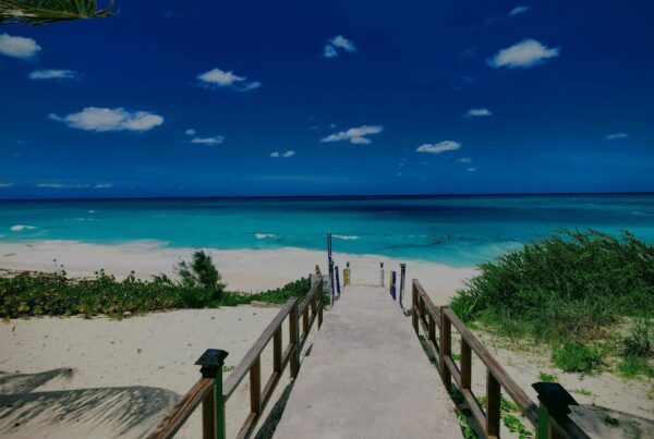 A wooden walkway with railings leads down to a sandy beach with turquoise water under a bright blue sky dotted with small white clouds. Green bushes line the sides of the path.