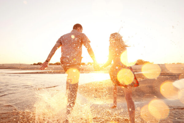 A couple holding hands runs through shallow water on a beach at sunset, splashing and surrounded by warm sunlight and lens flares—capture the joy of travel and maximize your PTO with unforgettable moments like these.