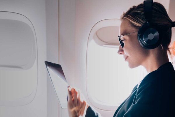 A woman wearing headphones and sunglasses sits in an airplane seat by the window, looking at a tablet. Bright daylight streams through, with an in-flight entertainment screen ahead—showcasing travel hacks for a smarter journey.