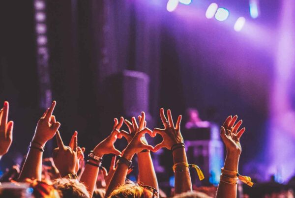 A lively Family Spring Break concert crowd raises their hands, some making heart shapes, under vibrant stage lights with a performer in the background. The scene bursts with energy and excitement.