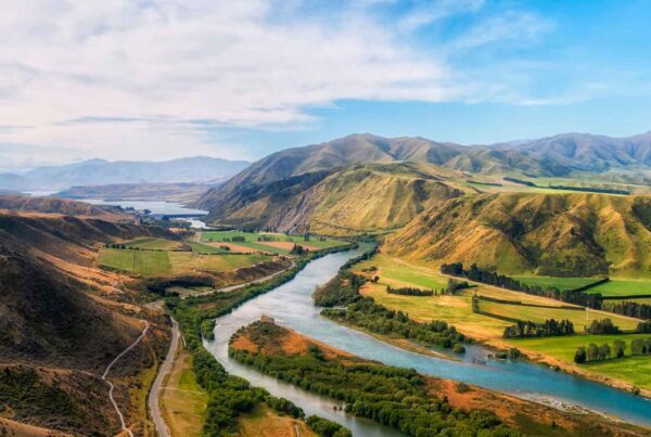 A winding river flows through a lush green valley, perfect for a Family Spring Break adventure, surrounded by rolling hills and distant mountains under a partly cloudy blue sky.