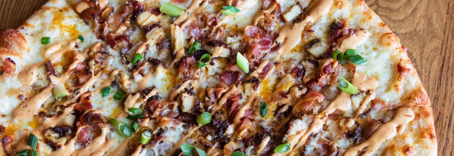 A close-up of a pizza topped with bacon, chicken, green onions, and a drizzle of creamy sauce on a wooden surface—perfect for foodies exploring Great Cities for Your Favorite Foods.