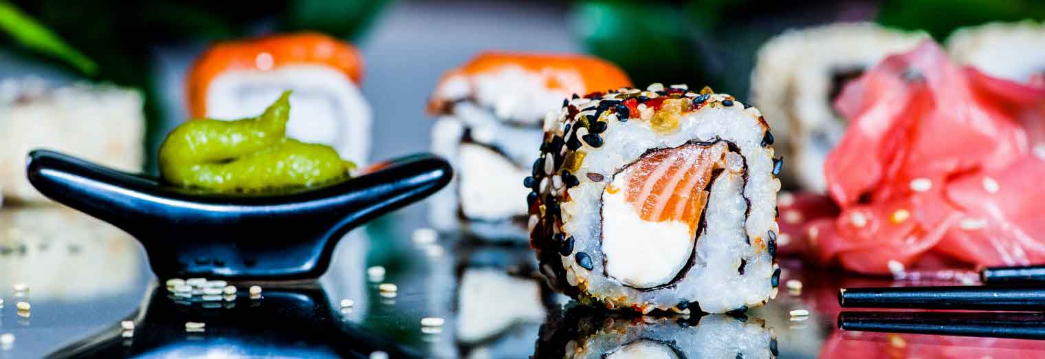 A close-up of assorted sushi rolls with salmon and cream cheese, garnished with sesame seeds, beside wasabi, pickled ginger, and chopsticks—perfect for exploring great cities for your favorite foods.