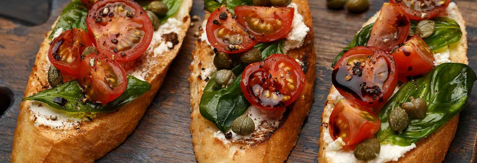 Three slices of toasted bread topped with ricotta cheese, fresh basil, juicy cherry tomatoes, capers, and cracked black pepper—just a taste of what you might find in the Great Cities for Your Favorite Foods.