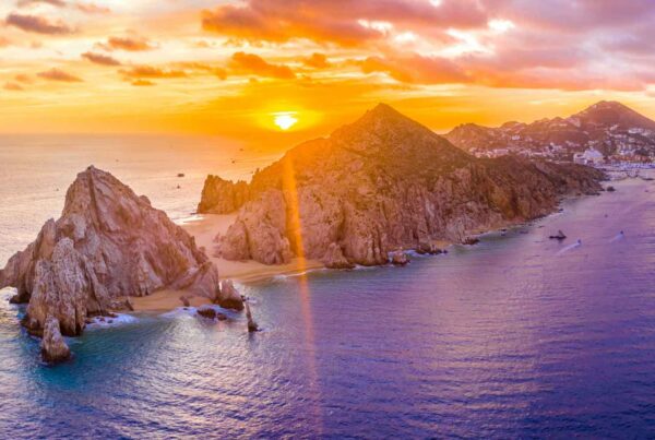 A vibrant sunset over rocky cliffs and clear blue waters at Land’s End, Cabo San Lucas, Mexico—perfect for Autumn Vacation Values—with mountains and the city visible in the background.