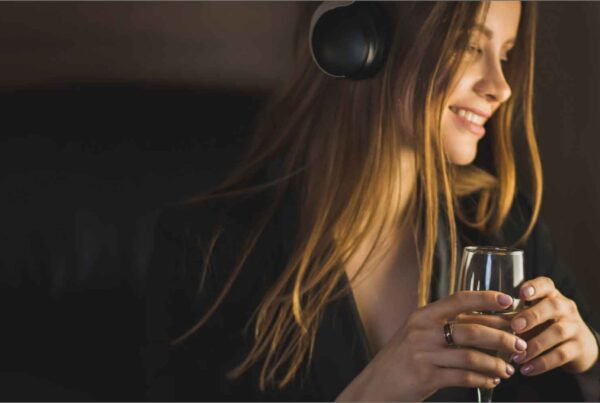 A woman with long hair, wearing headphones and a black outfit, sits by an airplane window smiling and holding a glass of champagne, savoring Autumn Vacation Values.
