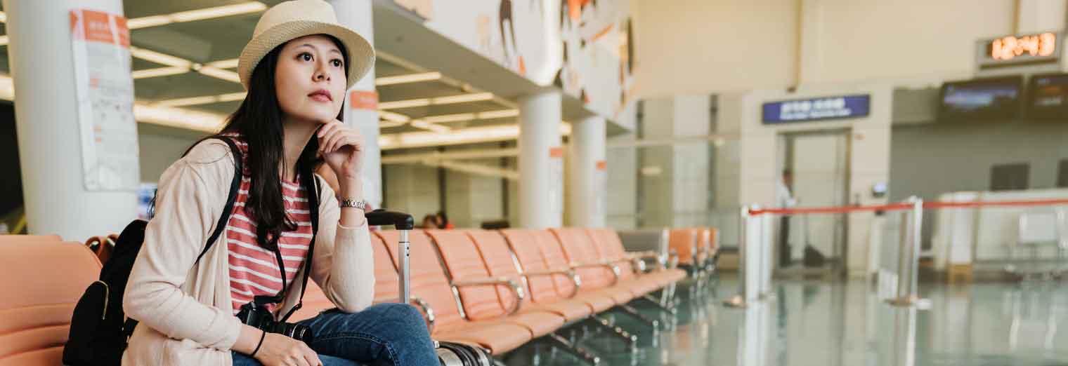 A woman wearing a hat and backpack sits on a bench in an empty airport terminal, looking thoughtful—perhaps reflecting on Autumn Vacation Values. Rows of orange seats and airport signs are visible in the background.