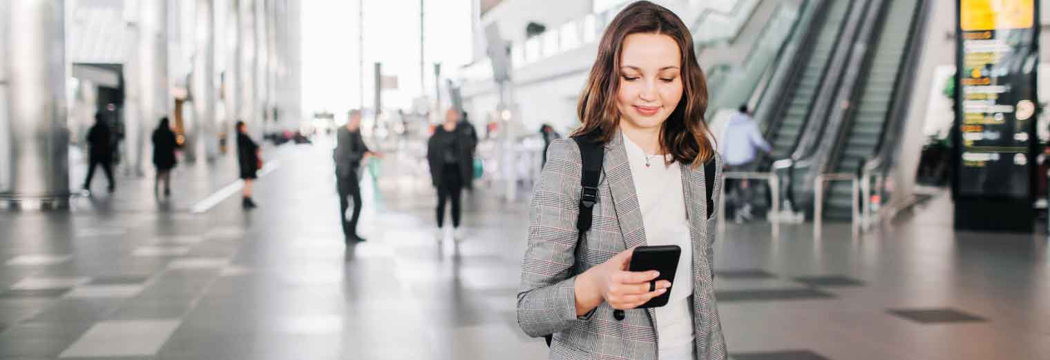 A young woman in a plaid blazer smiles at her smartphone while walking indoors, perhaps checking Autumn Vacation Values. She carries a backpack, with several people and an escalator in the modern, spacious building background.