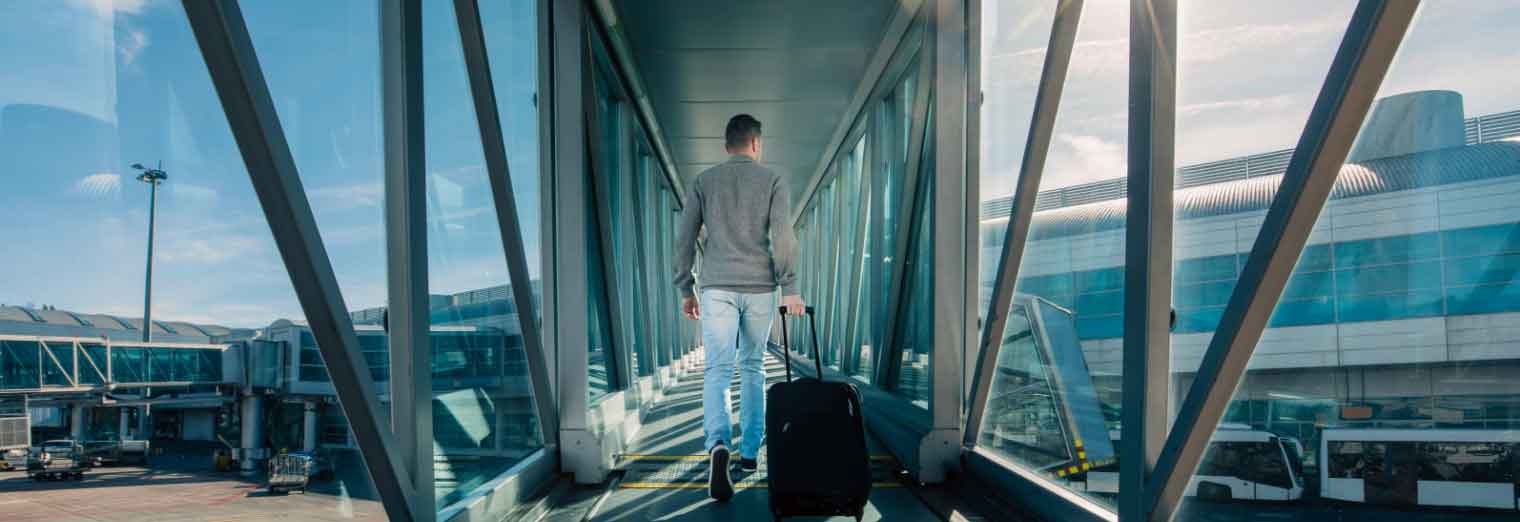 A person in casual clothes walks down an airport jet bridge, pulling a black suitcase, ready to enjoy Autumn Vacation Values, with terminal buildings and airplanes visible outside through glass walls.