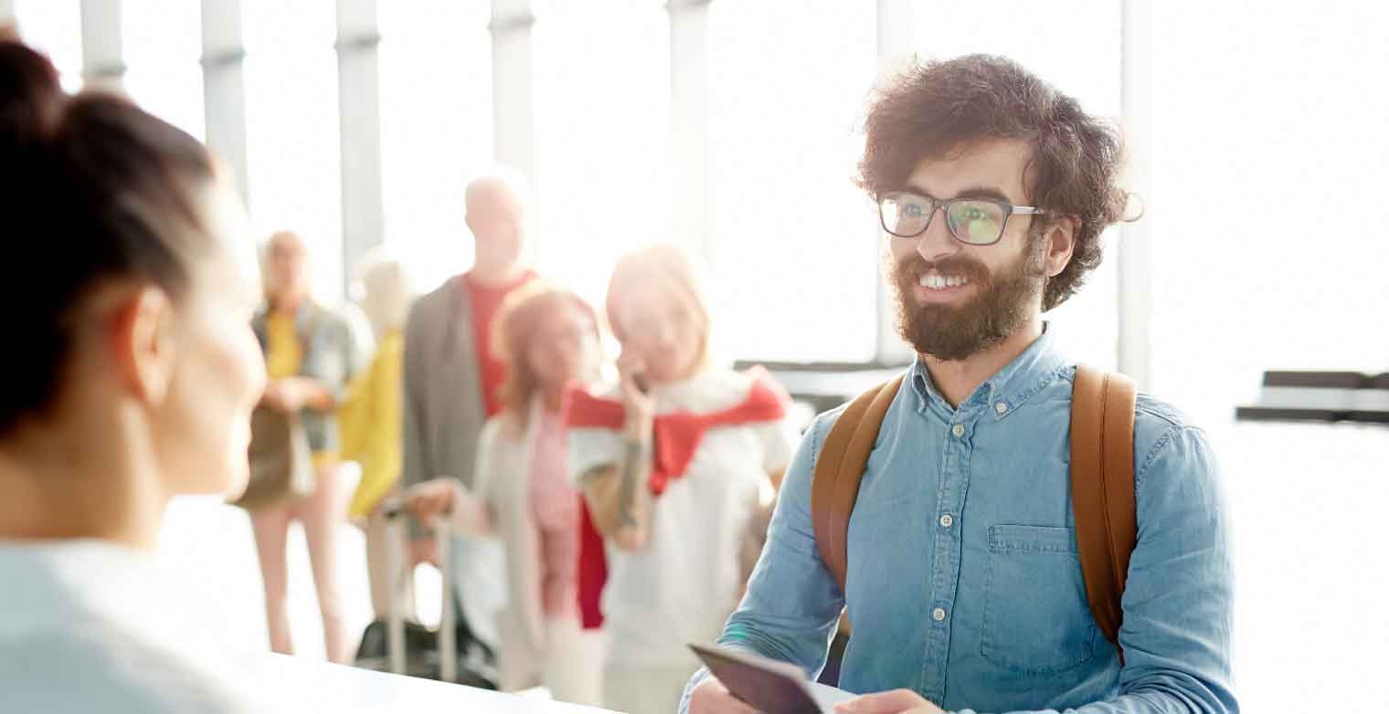 A smiling man with glasses and a backpack hands documents to an airline staff member at an airport counter, enjoying Autumn Vacation Values as people stand in line behind him with luggage.