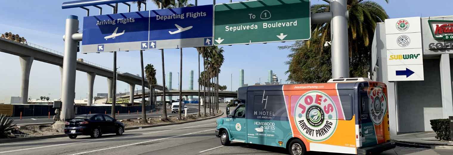 A colorful shuttle van drives past highway signs for arriving and departing flights and Sepulveda Boulevard at an airport, where palm trees line the road—perfect for travelers seeking Autumn Vacation Values. Cars and buildings fill the lively background.