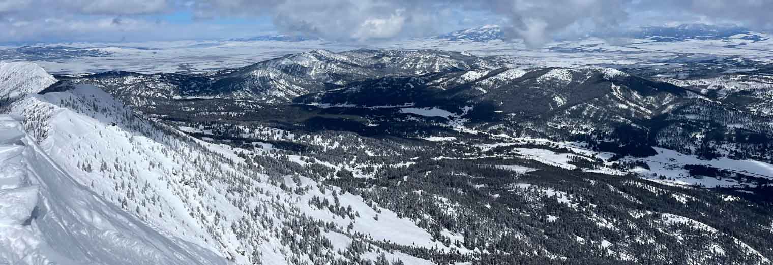 A panoramic view of snow-covered mountains and forested valleys under a partly cloudy sky, with patches of sunlight illuminating the landscape—perfect for discovering Autumn Vacation Values in nature’s serene beauty.