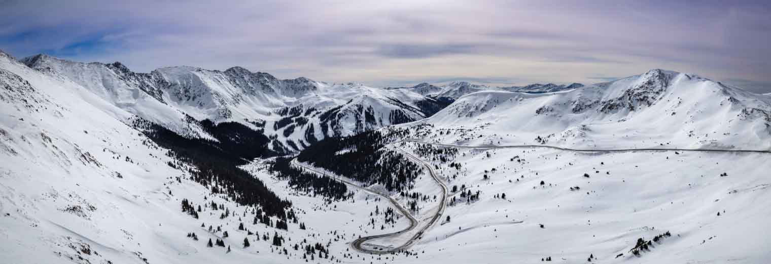 Snow-covered mountains under a cloudy sky with a winding road and scattered trees leading through the valley, surrounded by tall peaks—perfect for exploring Autumn Vacation Values.