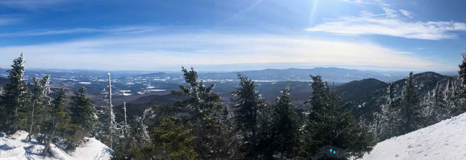 A panoramic view from a snowy mountain summit, overlooking frosted evergreen trees and a vast landscape of distant hills, valleys, and a blue sky with wispy clouds—a perfect scene to inspire your next Autumn Vacation Values adventure.