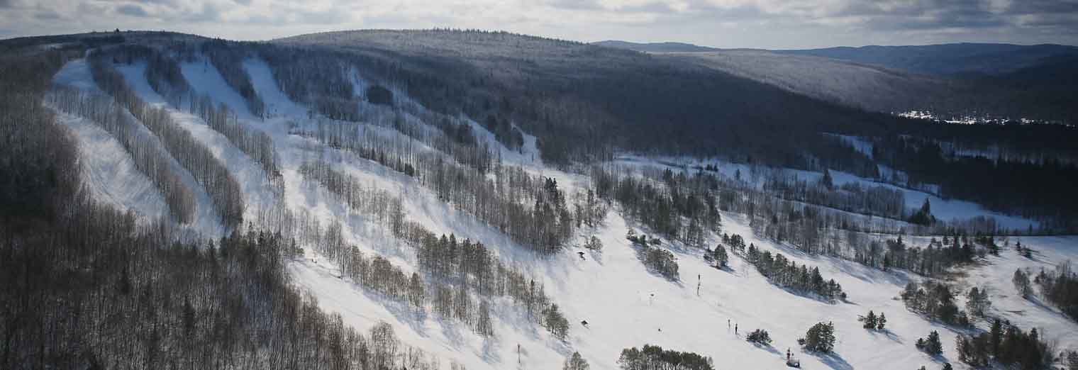 Aerial view of a snow-covered mountain with several ski trails winding through leafless trees under a cloudy sky, capturing the serene beauty and Autumn Vacation Values of an off-season escape.