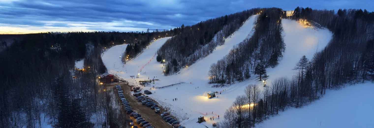 A snowy ski resort at dusk with lit ski slopes, trees on both sides, and a long parking lot lined with cars at the base. Small groups of people near the ski lifts enjoy Autumn Vacation Values on their winter getaway.
