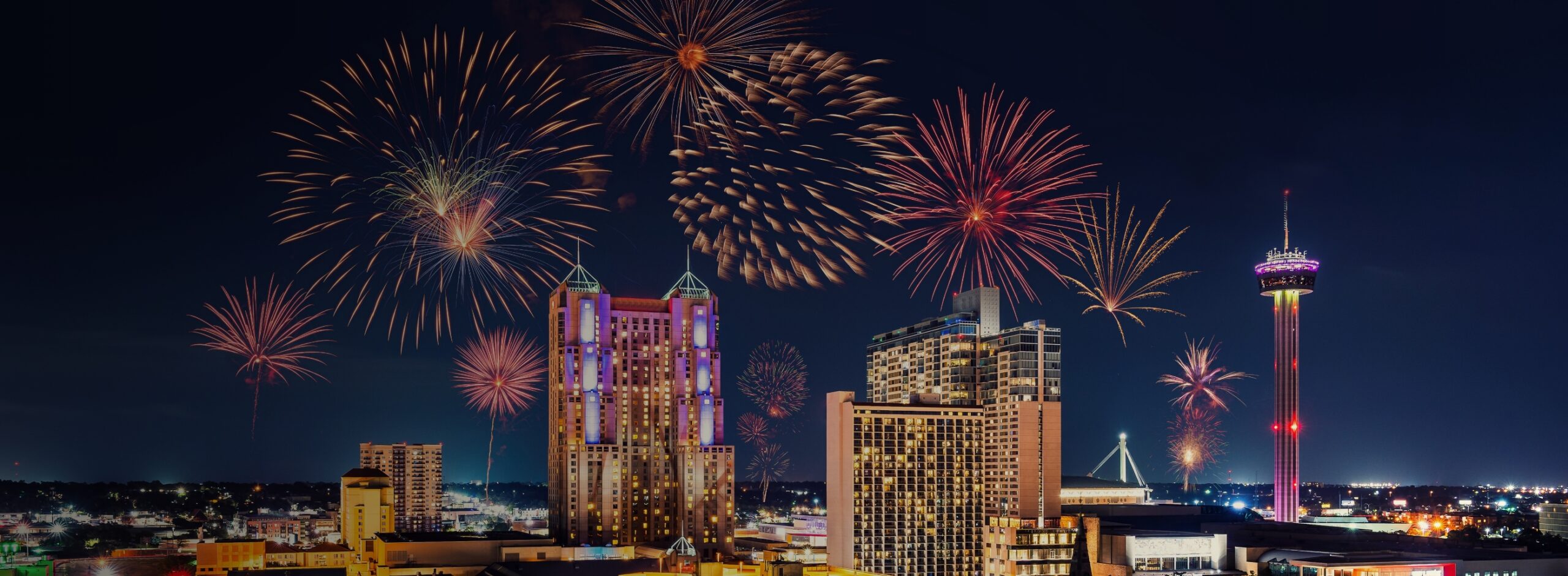 Colorful fireworks light up the night sky above a city skyline featuring tall buildings, a lit observation tower, and Joe's Airport Parking, creating a festive celebration atmosphere.