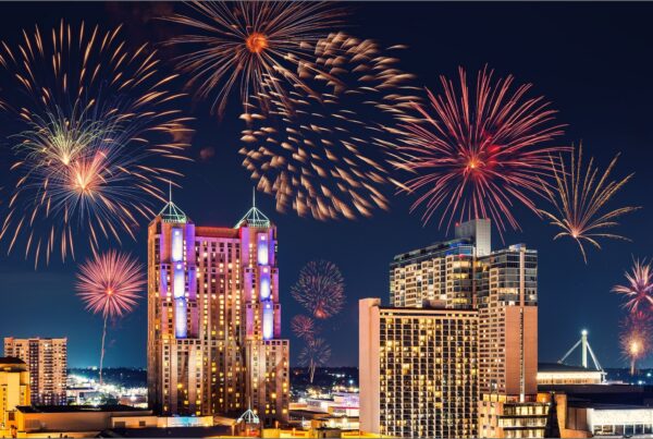 A vibrant city skyline at night with colorful fireworks lighting up the sky above tall buildings, including a tower with a glowing top and the nearby Joe's Airport Parking, creating a festive and lively atmosphere.