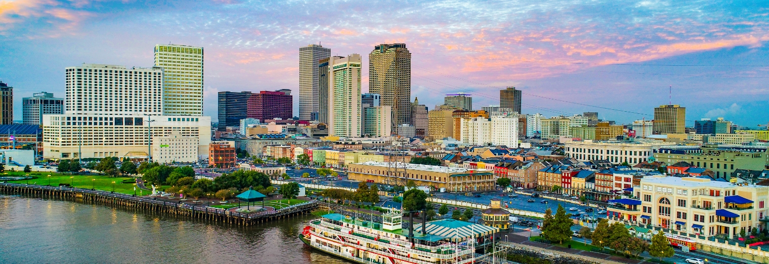 A colorful sunset sky over downtown New Orleans, showing tall modern buildings, historic French Quarter architecture, and a riverboat docked by the Mississippi River—just steps from conveniences like Joe's Airport Parking.