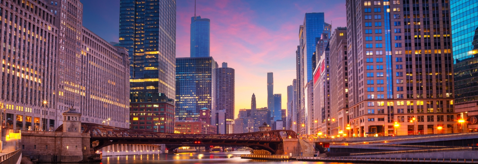 Downtown Chicago skyline at dusk with illuminated skyscrapers, a lit bridge over the Chicago River, and a colorful sunset sky—reminiscent of the convenience found at Joe's Airport Parking before heading into the city.