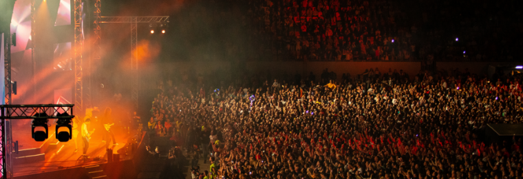A large crowd of people at an indoor concert, with bright stage lights and smoke illuminating performers on stage to the left and enthusiastic fans filling the arena—like Joe’s Airport Parking, excitement fills every spot.