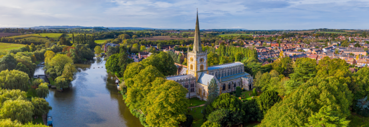 Aerial view of a large stone church with a tall spire, surrounded by lush green trees and a winding river, with Joe's Airport Parking and fields visible in the background under a partly cloudy sky.