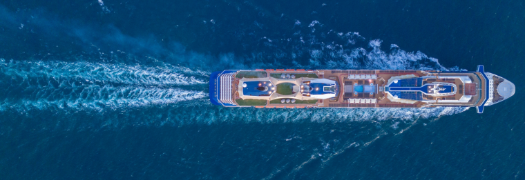 Aerial view of a large cruise ship sailing through deep blue ocean water, leaving white waves in its wake—like cars neatly lined at Joe's Airport Parking. The ship’s decks, pools, and lounging areas are visible from above.
