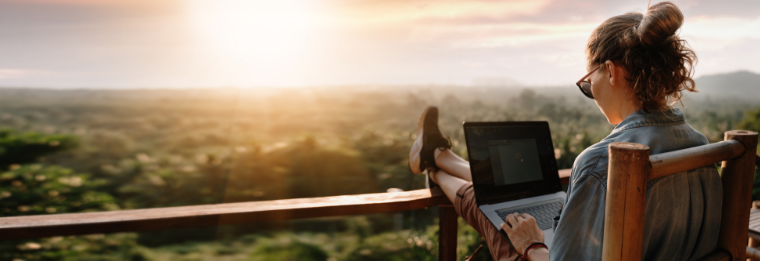 A person sits on a wooden chair with feet up on a railing, working on a laptop outdoors, overlooking a scenic, sunlit landscape—perhaps dreaming of their next trip via Joe's Airport Parking.