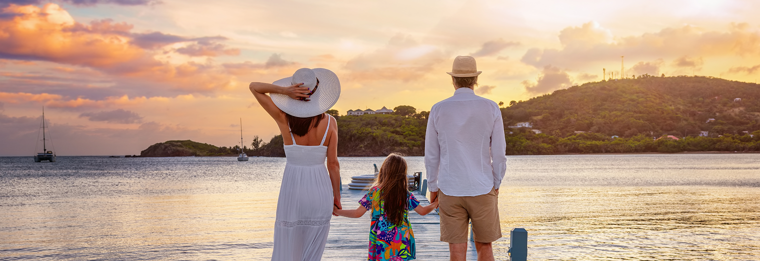 A woman, a man, and a young girl stand on a dock holding hands, looking at a sunset over a calm bay with sailboats and green hills in the distance—enjoying vacation time after parking their car at Joe's Airport Parking.