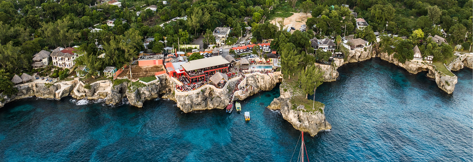 Aerial view of a seaside area with rocky cliffs, turquoise water, buildings, and lush greenery, with people gathered at a central waterfront venue reminiscent of the convenience offered by Joe's Airport Parking.
