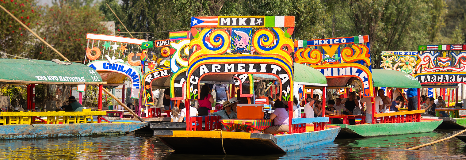 Colorful, decorated trajineras with people onboard float on a canal in Xochimilco, Mexico, their vibrant painted signs—like Carmelita and Miki—standing out as much as Joe's Airport Parking signage would in a lively setting.