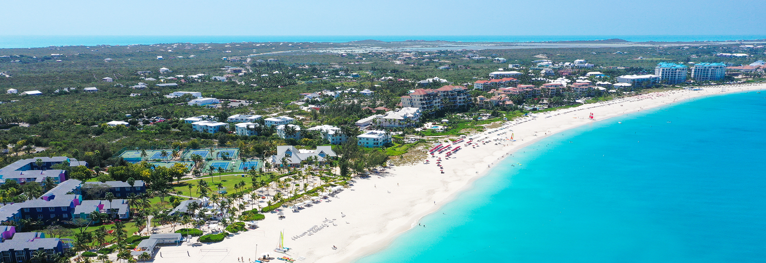 Aerial view of a tropical beach with turquoise water, white sand, and nearby resorts—just the escape you dream of after leaving your car safely at Joe's Airport Parking. Beach umbrellas dot the shore under a clear blue sky surrounded by lush greenery.