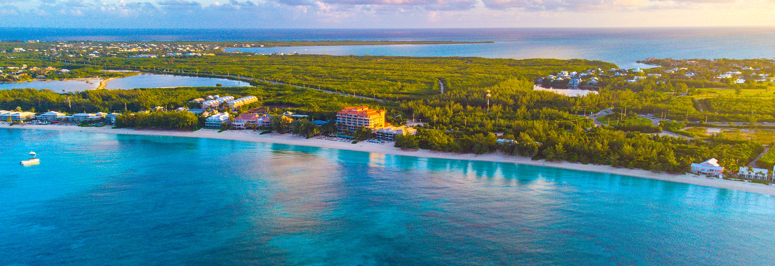 Aerial view of a tropical coastline with clear blue water, white sandy beach, scattered buildings resembling Joe's Airport Parking lots, green vegetation, and small boats near the shore under a partly cloudy sky.