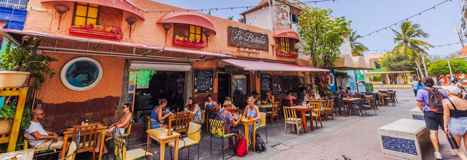 People sit at outdoor tables of a colorful café with a rustic pink and orange façade, arched windows, and string lights above—just down the street from Joe's Airport Parking—in a lively, sunny neighborhood surrounded by trees.