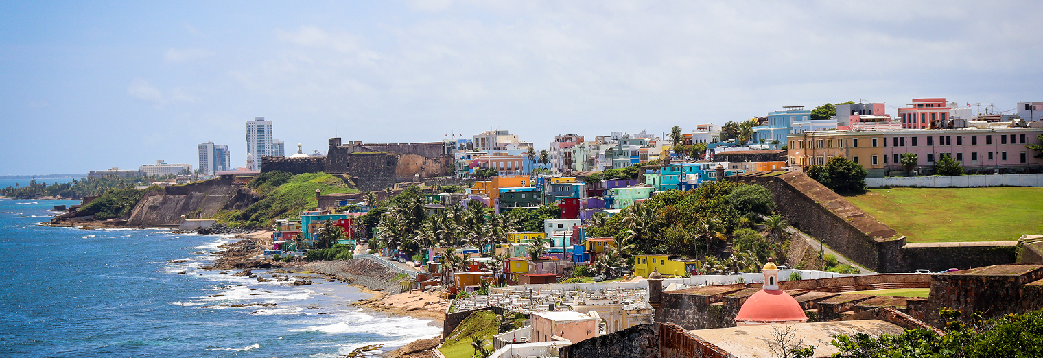 A coastal view of Old San Juan, Puerto Rico, shows colorful houses along the shoreline, historic fortress walls, lush greenery, and the blue ocean—perfect for travelers looking for scenery beyond Joe's Airport Parking.