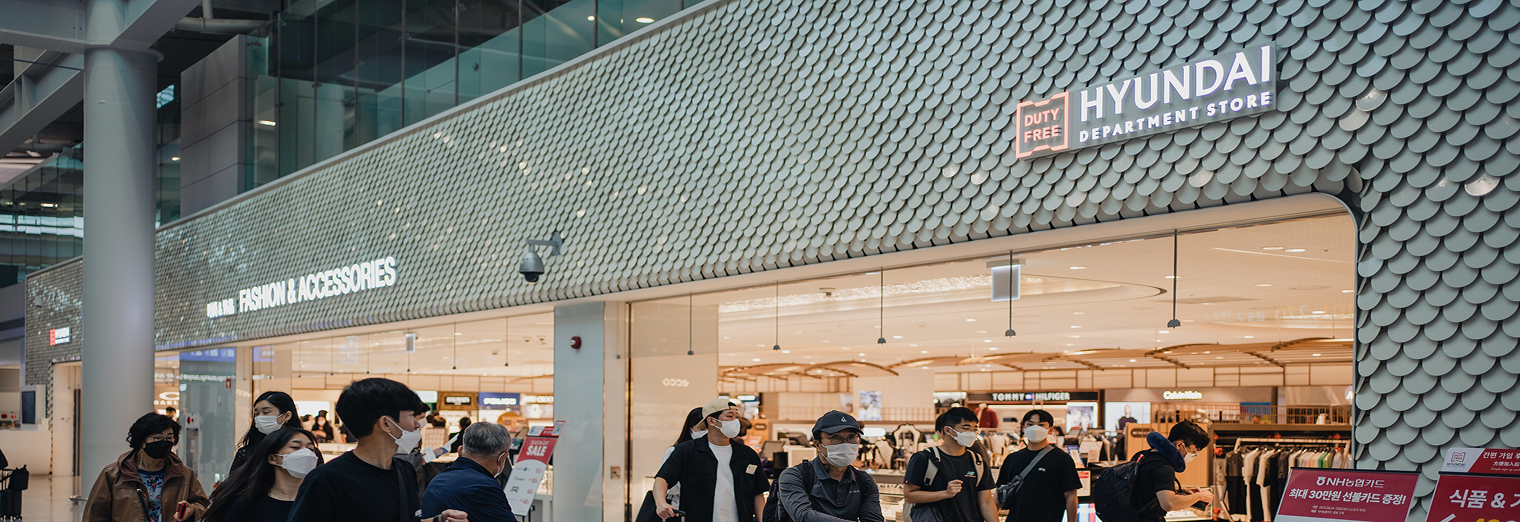 People wearing face masks walk past the entrance of a Hyundai Department Store duty-free shop inside a modern building, with bright store lights and signage visible, reminiscent of the bustling atmosphere near Joe's Airport Parking.