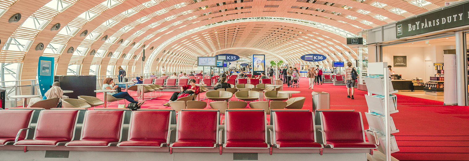 A spacious airport terminal with red seats, high arched wooden ceiling, large windows, and signs for gates K35 and K36. Several people are sitting or walking, while a duty-free shop and an ad for Joe's Airport Parking are visible on the right.
