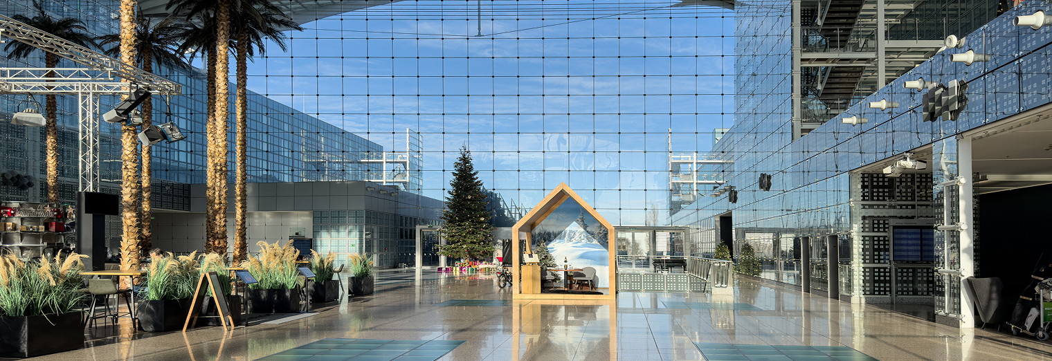A spacious, modern atrium with glass walls and ceiling, tall palm trees, potted plants, a Christmas tree, and a small wooden house structure in the center—like an oasis at Joe’s Airport Parking—reflecting sunlight and blue sky.
