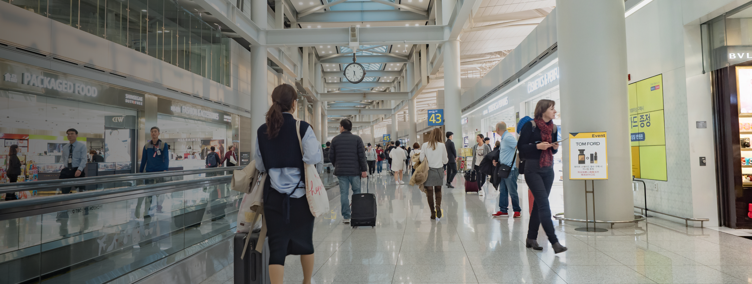 Busy airport terminal with people walking, pulling suitcases, and shopping. Large clock hangs from the ceiling, and signs for shops, gates, Joe's Airport Parking, and restaurants are visible. Bright lighting and modern architecture throughout.