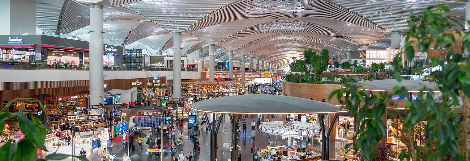 Wide view of a modern, bustling airport terminal with many travelers, shops, restaurants, and greenery beneath a curved ceiling—perfect for those arriving from Joe's Airport Parking.
