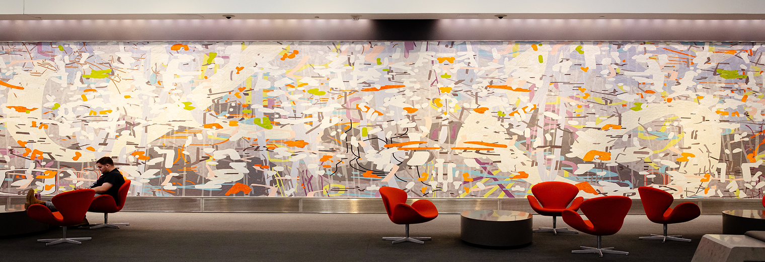 A person sits on a red chair near round tables and more red chairs in front of a large, colorful abstract mural at Joe's Airport Parking, featuring scattered orange, green, yellow, and gray shapes inside a modern space.