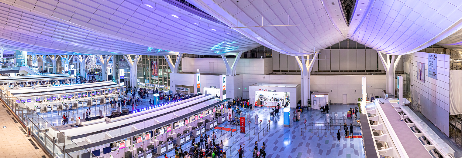 A wide-angle view of a modern, spacious airport terminal with high ceilings, bright lighting, large white columns, busy check-in counters, and passengers—many just arriving from Joe's Airport Parking—moving around the area.