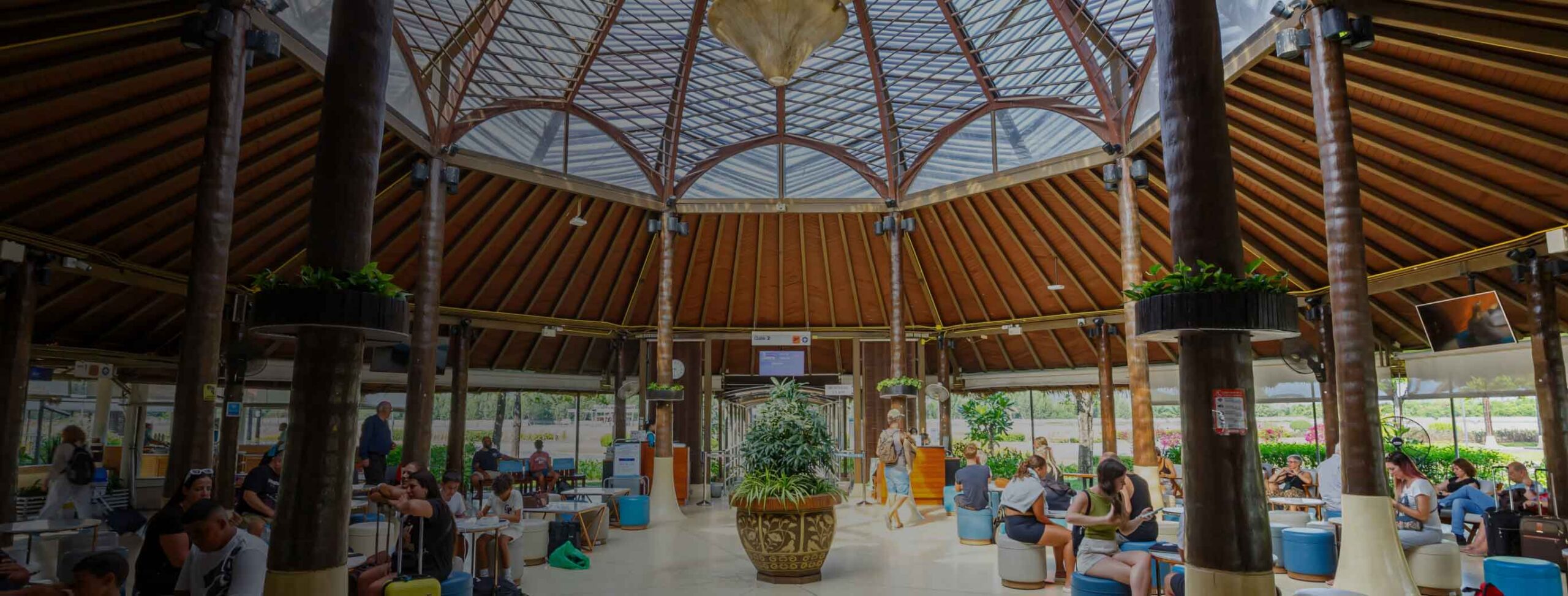 A spacious, circular indoor area with a high, wooden, dome-shaped ceiling and glass panels. Like the lounge at Joe's Airport Parking, people sit at tables around a central plant, enjoying food and drinks in a bright, airy setting.