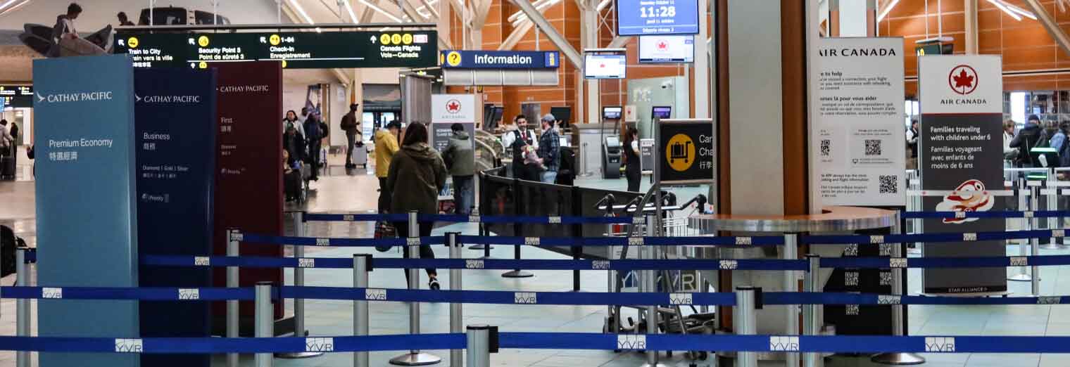 Passengers stand in line at an airport check-in area with signs for Cathay Pacific and Air Canada. Blue queue barriers guide travelers, while overhead signs display flight details—many arrive after using Joe's Airport Parking.