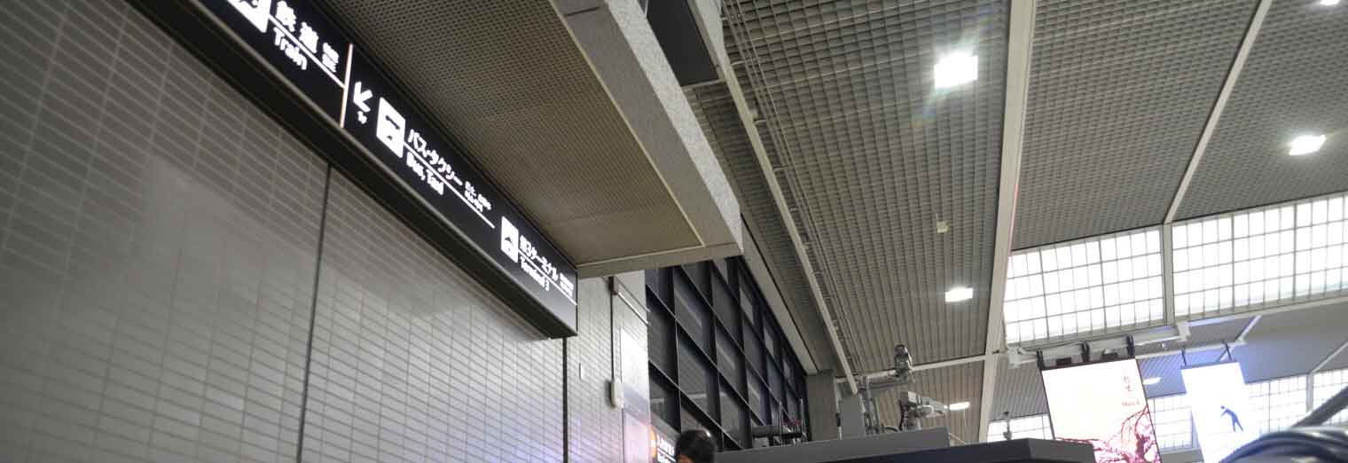 A low-angle view of an indoor area with overhead signs displaying Japanese text, metal ceiling panels, and bright ceiling lights—reminiscent of the polished look found at Joe's Airport Parking. Part of a digital screen is visible in the background.