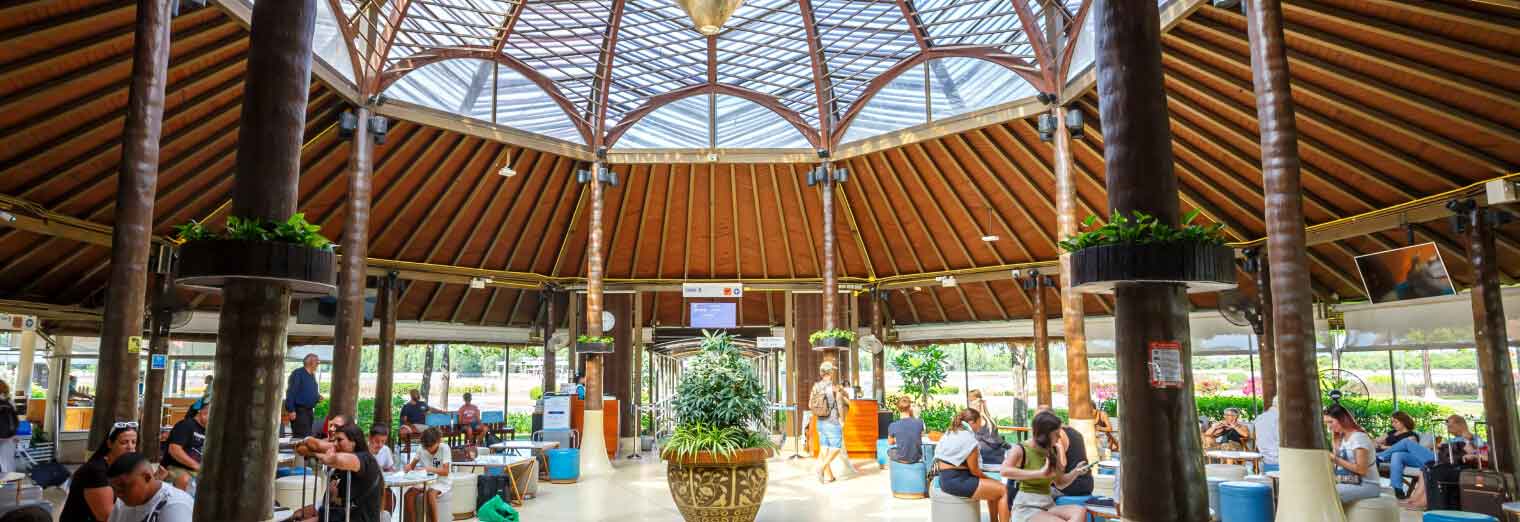 Travelers sit and wait in a spacious, circular airport terminal with a high, wooden ceiling and abundant natural light. Some discuss their plans while others check phones for updates from Joe's Airport Parking before their next flight.