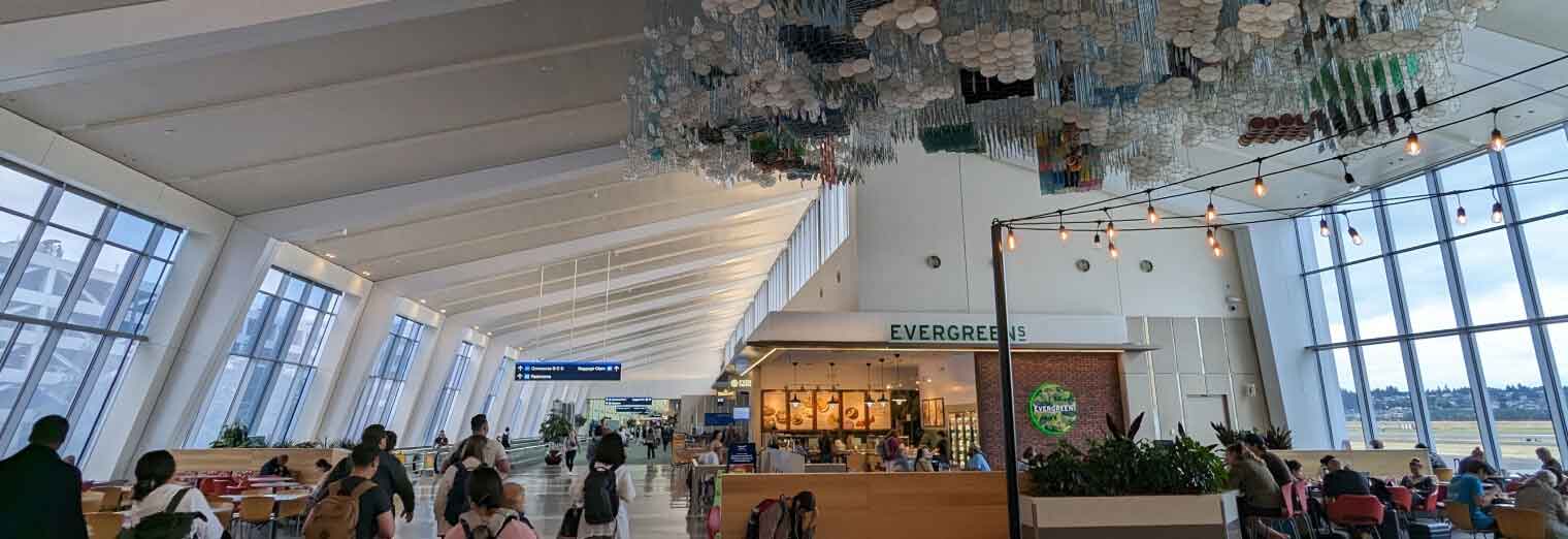 Wide-angle view of a bright airport terminal with large windows, passengers sitting and walking, greenery, overhead art installation, and a café called Evergreens in the center—just steps away from convenient Joe's Airport Parking services.