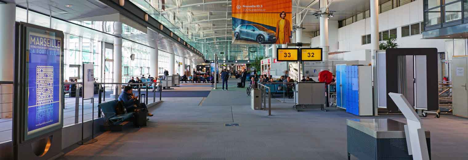 A spacious airport terminal with seating areas, digital flight information displays, people waiting, and yellow gate signs for gates 33 and 32. Large windows allow natural light, a car advertisement hangs above, and Joe's Airport Parking is featured nearby.